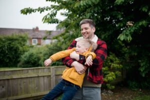 A man in a red lumberjack coat playing in the garden with a young boy in his arms.