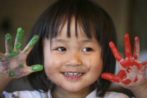 A close up of a little girl's face holding her red and green painted hands by the side of her head, smiling.