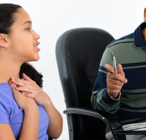 A teen girl sat looking and talking to a male adult who is mostly off camera with her hands resting on her upper chest
