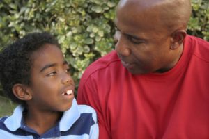 A young boy smiling with a male adult looking at him outdoors,