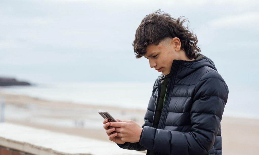 lone teenage boy looking at his phone screen while leaning against the wall on the seafront